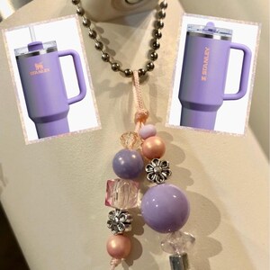 May include: A purple stainless steel travel mug with a handle and a straw, with the word "STANLEY" printed on the side. The mug is attached to a chain with a charm made of purple, pink, and clear beads.