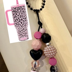 May include: A tumbler with a pink and black leopard print pattern, a pink handle, and a straw, paired with a decorative beaded accessory featuring black, pink, and light pink beads in various shapes, including a heart and a textured sphere.