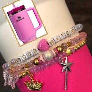 May include: Pink and gold charm bracelets with a pink heart, a white pearl, a gold crown, and a silver star wand charm. The bracelets spell out "GOOD WITCH".