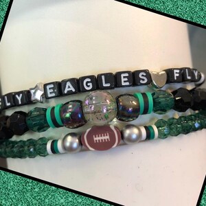 May include: Three beaded bracelets. The top bracelet spells "FLY EAGLES FLY" with black letter beads. The other bracelets feature green, black, and clear beads, with a football-shaped bead on the bottom bracelet.