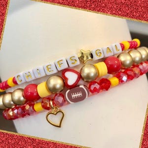 May include: Three beaded bracelets in red, gold, and yellow. One bracelet spells "CHIEFS GAL" with letter beads and a gold star charm. Other bracelets feature heart, football, and gold heart charms. The bracelets are on a white background.