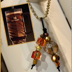 May include: A Stanley travel mug with a brown and tan tiger stripe design. The mug has a black handle and the Stanley logo. A gold beaded charm with orange and gold beads and a star charm hangs from a gold chain.