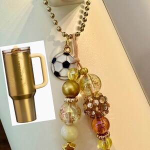 May include: A gold-colored Stanley tumbler with a handle is shown next to a gold-toned necklace. The necklace features a soccer ball charm, gold beads, and a star charm. The tumbler has a lid and a handle.