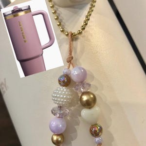 May include: A pink Stanley tumbler with a handle and gold accents, next to a beaded charm with gold, white, and iridescent beads. The tumbler has the brand name "STANLEY" printed on it.