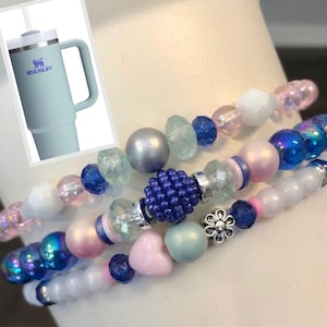 May include: Three beaded bracelets with a variety of beads in shades of pink, blue, white, and iridescent. A light blue Stanley tumbler with a handle and the Stanley logo is in the upper left corner.