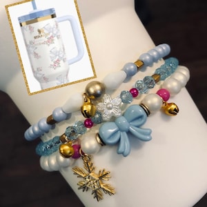 May include: Four blue, white, and pink beaded bracelets with gold charms, including a snowflake, bells, and bows. The bracelets are stacked on a white surface.