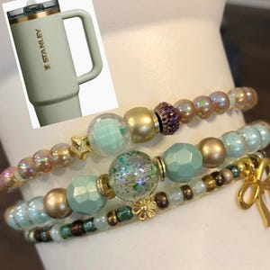 May include: A light green Stanley tumbler with a handle and gold accents is next to three beaded bracelets. The bracelets feature iridescent, gold, and mint green beads, with a gold bow charm.