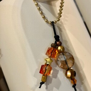 May include: A gold-coloured chain with a black cord and decorative beads. The beads include orange and clear faceted cubes, a gold star, a large round clear bead, and gold and brown spherical beads. The chain is attached to a white surface.
