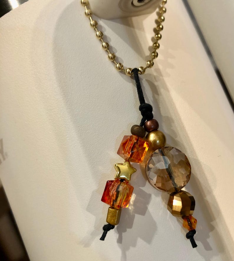 May include: A gold-coloured chain with a black cord and decorative beads. The beads include orange and clear faceted cubes, a gold star, a large round clear bead, and gold and brown spherical beads. The chain is attached to a white surface.