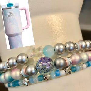 May include: A set of three beaded bracelets with silver, blue, and pink beads, and star charms. A large insulated tumbler with a pink handle and a blue and white marbled design is in the background.