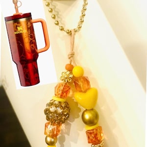 May include: A red and gold Stanley tumbler with a handle and pineapple-shaped straw topper. A beaded charm necklace with yellow, gold, and orange beads, including a heart-shaped bead and a flower charm, hangs nearby.