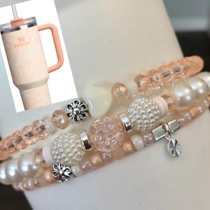 May include: A stack of three beaded bracelets in shades of pink, white, and silver. The bracelets feature various bead types, including pearls, clear beads, and decorative silver accents. A Stanley tumbler with a peach handle and floral design is in the background.