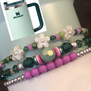 May include: Four beaded bracelets with green and pink beads, white flower charms, and silver beads. The bracelets are wrapped around a light green Stanley travel mug.