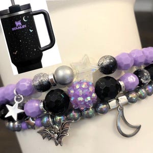 May include: Black Stanley tumbler with a handle, featuring a starry night design. Three beaded bracelets with purple, black, silver, and iridescent beads. Charms include a star, a bat, and a crescent moon.