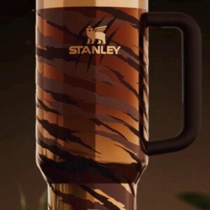 May include: A large Stanley tumbler with a black handle and lid. The tumbler features a tiger stripe pattern in shades of brown and gold. The Stanley logo is visible on the front.
