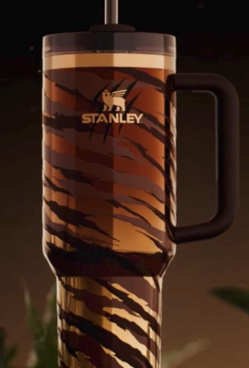 May include: A large Stanley tumbler with a black handle and lid. The tumbler features a tiger stripe pattern in shades of brown and gold. The Stanley logo is visible on the front.