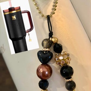 May include: Black insulated tumbler with a handle and a gold logo, next to a beaded charm with a black heart, gold, and black beads. The tumbler has a red lid and straw.
