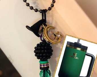 May include: A black beaded charm with a green and purple accent hangs from a black chain. The charm is attached to a gold crescent moon and star charm. A black witch's hat charm is also attached to the chain. The charm is hanging from a Stanley green travel mug with a gold lid and handle.