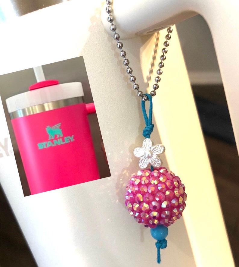 May include: A pink and silver charm with a flower and a sparkly ball hangs from a silver chain. The charm is attached to a pink Stanley water bottle.