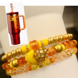 May include: Three beaded bracelets in shades of gold, yellow, and orange. The bracelets feature various bead shapes, including round, square, and faceted. A red and gold insulated tumbler with a handle and straw is also visible.