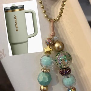 May include: A light green Stanley tumbler with a handle and gold accents. The tumbler is next to a beaded charm with gold, iridescent, and turquoise beads, and a gold star charm. A gold chain is also visible.