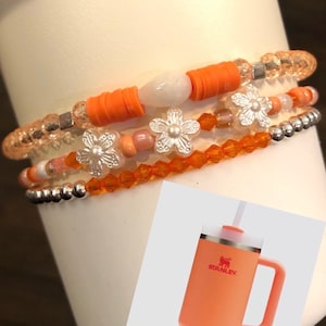 May include: A set of four bracelets with orange and white beads, featuring white flower charms. The bracelets are stacked on a white surface.