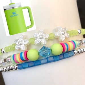 May include: Three beaded bracelets with a variety of colorful beads. One bracelet features white flower-shaped beads with pearl centers. Another has rainbow-colored disc beads and a light blue square bead. A green Stanley tumbler is in the background.
