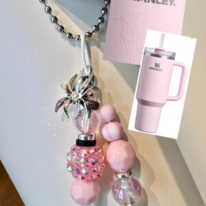 May include: A decorative charm featuring a silver spider pendant and various pink beads, including faceted, iridescent, and solid spheres. The charm is attached to a silver ball chain and is displayed alongside a pink card with the Stanley logo and a graphic of a pink tumbler.