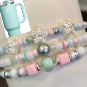 May include: Four beaded bracelets with silver flower charms, pink, white, and light blue beads. The bracelets are stacked on top of each other.