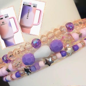 May include: A set of four beaded bracelets with pink, purple, and clear beads. The bracelets feature silver star charms and a white bead. The bracelets are stacked on top of each other.