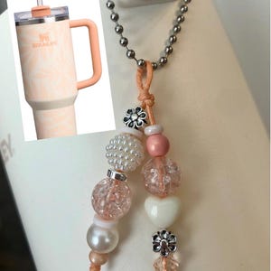 May include: A Stanley tumbler with a peach-colored handle and lid, featuring a floral pattern. A beaded charm with pink, white, and clear beads, a white heart, and silver flower accents hangs from a chain.