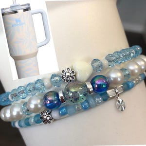 May include: A light blue Stanley tumbler with a floral design and a set of three beaded bracelets. The bracelets feature a mix of blue, white, and iridescent beads, with silver accents and charms. The tumbler has a handle and a straw.