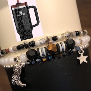 May include: Three layered bracelets with black, white, and gold beads, charms, and stars. The bracelets are on a black background with a silver boot charm hanging from the bottom.