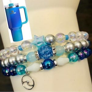 May include: A blue and teal gradient tumbler with a handle and straw is shown next to three beaded bracelets. The bracelets feature a variety of blue, silver, and clear beads, including a star-shaped bead and a charm with a wave design.