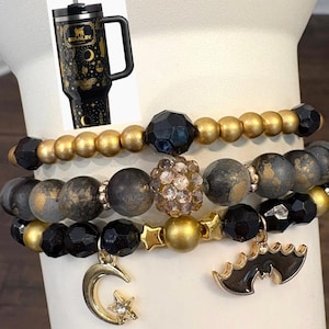 May include: A set of three beaded bracelets featuring gold, matte black, and textured grey beads. One bracelet includes a gold crescent moon charm with a star, and another features a black bat charm. A black tumbler with gold celestial motifs is shown.
