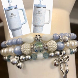 May include: Two light blue Stanley tumblers with floral patterns and handles are displayed above three beaded bracelets. The bracelets feature pearls, silver beads, and charms. The bracelets have blue, white, and silver beads.