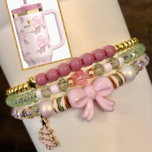 May include: A set of four pink and green beaded bracelets with a pink bow charm and a pink Christmas tree charm. The bracelets are stacked on top of each other and have gold beads.