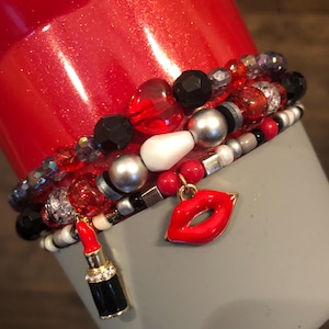 May include: A set of red, black, white, and silver beaded bracelets with a red lip charm and a red lipstick charm.