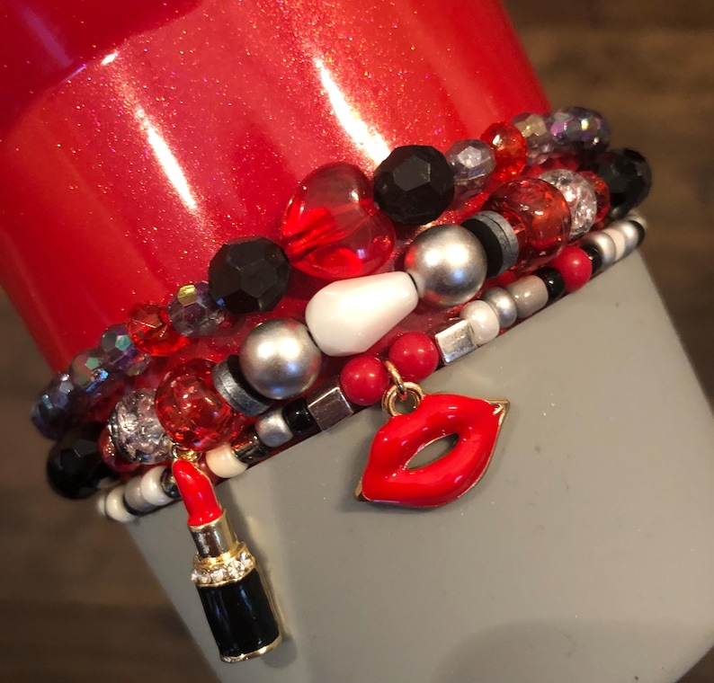 May include: A set of red, black, white, and silver beaded bracelets with a red lip charm and a red lipstick charm.