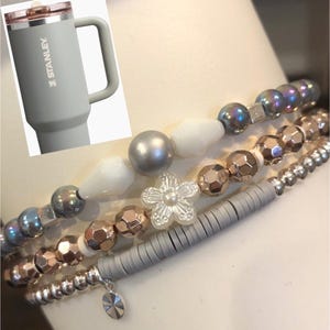 May include: A set of four bracelets with various beads, including silver, white, rose gold, and iridescent beads. The bracelets are stacked on top of each other and feature a white flower charm.
