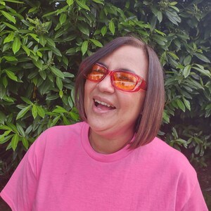 May include: A woman wearing a pink t-shirt and orange sunglasses laughs while standing in front of a green leafy background.