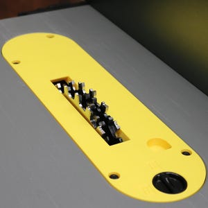 May include: A close-up of a table saw blade. The blade is black and silver, and is partially visible through a yellow safety guard. The table surface is gray.