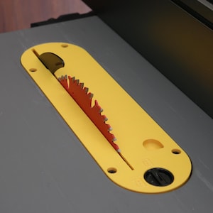 May include: A close-up of a table saw blade. The blade is red and has sharp teeth. It is partially covered by a yellow blade guard. The saw table is gray.