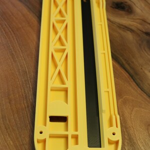 May include: A yellow plastic tool with a black insert, likely for woodworking. The tool has a rectangular shape with rounded ends and a series of geometric cutouts and a circular feature with a smiley face design.