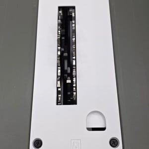 May include: A white table saw throat plate with a long rectangular center slot, a smaller semi-circular cutout, and a black circular locking knob at the bottom. The plate features two black screws and two small icons indicating locking positions.