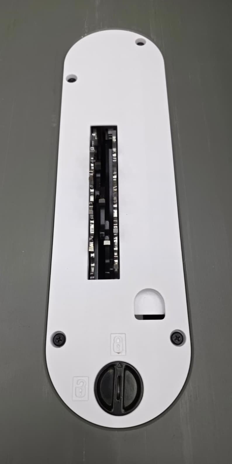 May include: A white table saw throat plate with a long rectangular center slot, a smaller semi-circular cutout, and a black circular locking knob at the bottom. The plate features two black screws and two small icons indicating locking positions.