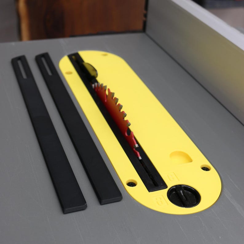 May include: A table saw blade with a bright yellow blade guard and two black anti-kickback pawls. The saw blade is red with sharp teeth. The items are on a gray surface.
