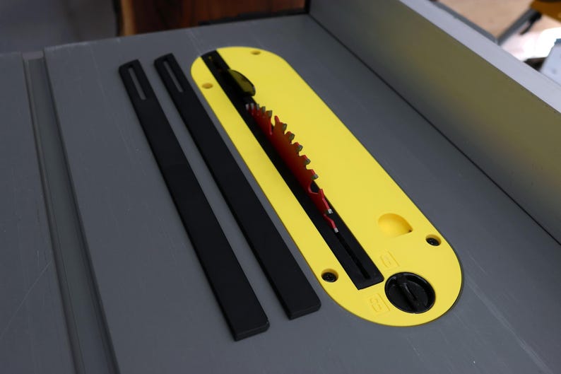 May include: A table saw insert plate in bright yellow with a central slot for a red circular saw blade. Two long, narrow black rectangular strips are positioned parallel to the insert plate on the gray surface of the table saw.