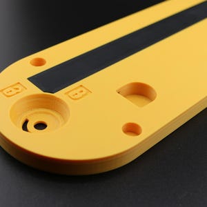 May include: Close-up of a yellow, oblong object with a black stripe and various circular cutouts. The object has a matte finish and appears to be made of plastic or a similar material. The background is a dark gray.