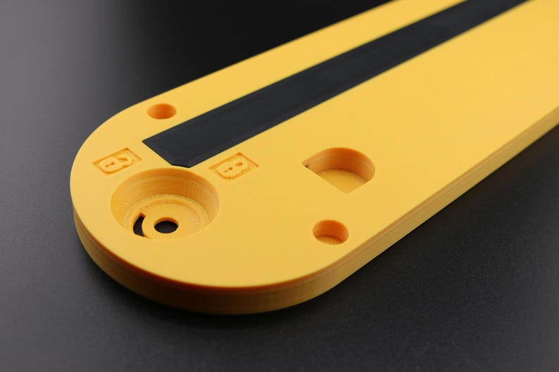 May include: Close-up of a yellow, oblong object with a black stripe and various circular cutouts. The object has a matte finish and appears to be made of plastic or a similar material. The background is a dark gray.